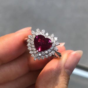 May include: A silver-toned ring featuring a large, heart-shaped, deep red gemstone at the center. The central stone is surrounded by a double halo of small, round, clear white diamonds set in a prong setting on a polished metal band.