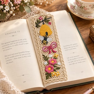 Può includere: Un segnalibro rettangolare in pizzo color crema con ricami dettagliati di un alveare giallo, un'ape nera e gialla, un fiocco rosa e un gruppo di fiori rosa e gialli con foglie verdi, posizionato all'interno di un libro aperto.