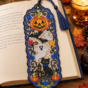 May include: An embroidered Halloween bookmark featuring a black lace-style border with blue accents. The design includes a jack-o'-lantern, a white ghost, a black cat, a haunted house, bats, a crescent moon, and colorful autumn leaves with a blue tassel.