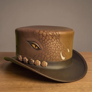 Puede incluir: Un sombrero de copa marrón con un patrón detallado de escamas de reptil y un gráfico de un ojo amarillo prominente en el lateral. El sombrero incluye una banda marrón oscuro adornada con una fila de pequeños adornos circulares.