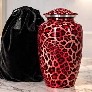 May include: A cremation urn featuring a vibrant red and black leopard print pattern with a metallic silver rim and a matching lid, accompanied by a black velvet drawstring bag with decorative tassels and braided cord detailing.