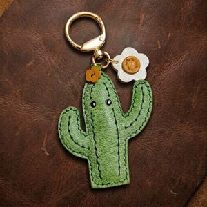 May include: A green leather cactus keyring featuring a gold-tone metal ring, a small orange flower accent on top, and a white flower charm with a yellow centre attached to the side. The cactus has two arms and two small black dot eyes.