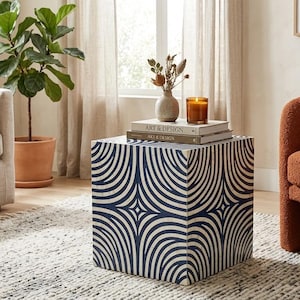 May include: A square side table featuring a repeating navy blue and white curved geometric pattern. The table surface holds two stacked books titled Art & Design, a small beige vase with dried botanicals, and a glass candle with a lit wick.