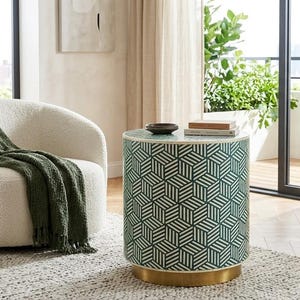 May include: A cylindrical side table featuring a green and white geometric cube pattern with a gold-toned base, positioned next to a cream-colored textured armchair draped with a dark green fringed throw blanket.