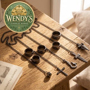 May include: A set of five dark metal rings connected by thin chains to various charms, including a small sphere, a cross, a hook, a sword, and a pointed arrowhead. The rings feature textured bands and are arranged in a row on a wooden surface.