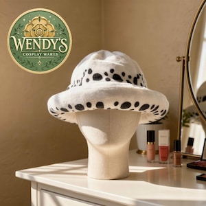 May include: A white bucket hat made of soft, plush material featuring a repeating pattern of irregular black spots. The hat is displayed on a neutral-coloured mannequin head. A circular logo in the upper left corner reads Wendy's Cosplay Wares.