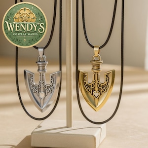 Puede incluir: Dos colgantes en forma de flecha colgados de cordones negros, uno en metal plateado y otro en dorado. Cada colgante presenta un intrincado diseño recortado con una pequeña forma de corazón en el centro. Un logo circular dice WENDY'S COSPLAY WARES.