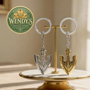 May include: Two metal arrow-shaped keychains hanging from a gold-toned display stand. One keychain is silver-coloured and the other is gold-coloured. Both feature intricate engraved patterns and a heart-shaped cutout. A circular logo reads WENDY'S COSPLAY WARES.