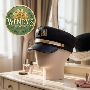 Pode incluir: Um boné de estilo militar preto com uma faixa dourada na frente, um pequeno botão dourado na lateral e um emblema retangular dourado exibindo um símbolo de mão. O boné é exibido em uma cabeça de manequim de tom neutro.