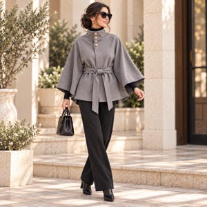 May include: A grey cape-style jacket with a high collar, light-colored buttons, and a matching fabric tie-waist belt. The garment is worn over a black long-sleeved top and black trousers, paired with black pointed-toe boots and a black handbag.
