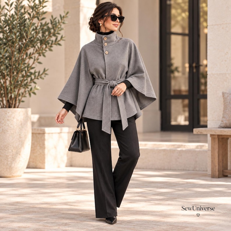 May include: A grey wool-blend cape coat featuring a high collar with three light-colored buttons, a matching tie-waist belt, and wide sleeves. The garment is worn over a black long-sleeved top and black trousers, paired with a black handbag.