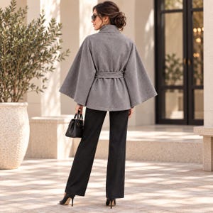 May include: A grey wool-blend cape coat featuring a high collar, wide flared sleeves, and a matching tie belt at the waist, paired with straight-leg black trousers and a small black structured handbag with top handles and gold-tone hardware.