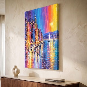 May include: A rectangular canvas wall art piece featuring a vibrant, textured oil painting of a city waterfront at sunset. The scene depicts colorful buildings, street lamps, and figures with umbrellas along a path, with sailboats on the water.