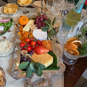 May include: A large, ornate silver serving tray with intricate embossed edges and two handles, holding an assortment of cheeses, red grapes, a small orange pumpkin, cherry tomatoes, dried apricots, a pear, and fresh green sage leaves.