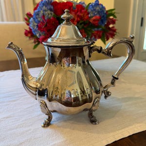 May include: A polished silver-plated teapot featuring a curved spout, a decorative lid with a finial, and an ornate handle. The body of the teapot has a rounded, bulbous shape supported by three detailed feet, reflecting the surrounding environment.