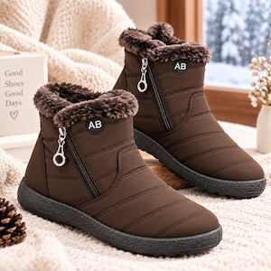 May include: A pair of brown ankle boots featuring a quilted exterior, a side zip closure with a circular pull, and a dark brown faux fur lining at the collar. Each boot displays a circular patch with the letters AB on the outer side.