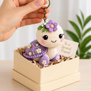 May include: A crocheted turtle keychain featuring a cream-colored head with pink cheeks, black eyes, and a purple shell decorated with small white flowers and green leaves. A purple flower sits on its head. A card reads To a Special Friend.
