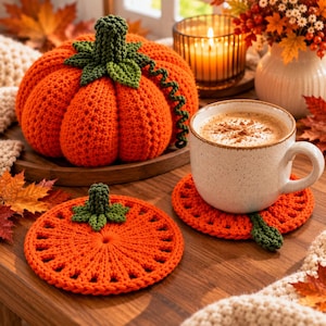 Puede incluir: Una calabaza de ganchillo naranja con tallo verde y zarcillo, acompañada de dos posavasos planos a juego con forma de calabaza. Uno está vacío mientras que el otro sostiene una taza de cerámica moteada con una bebida cremosa y canela.