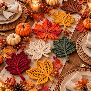 May include: A collection of crocheted leaf-shaped coasters in various colors including red, orange, yellow, cream, and dark green, arranged on a table runner alongside small pumpkins, berries, and place settings.