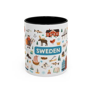 Pode incluir: Uma caneca de cerâmica branca com interior preto, apresentando um padrão colorido de ilustrações com temática sueca, incluindo um viking, um alce, uma casa vermelha, um navio e a palavra SWEDEN impressa numa faixa azul retangular.