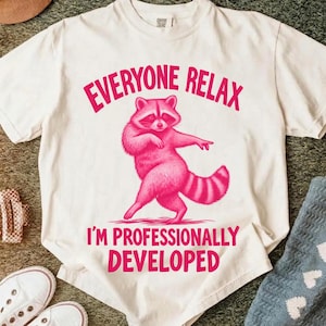 May include: A cream-coloured short-sleeve t-shirt featuring a graphic of a pink raccoon in a dancing pose. The text "EVERYONE RELAX" is printed in pink above the raccoon, and the text "I'M PROFESSIONALLY DEVELOPED" is printed in pink below it.