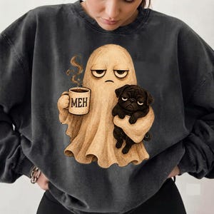 Könnte beinhalten: Ein anthrazitfarbenes Sweatshirt mit Rundhalsausschnitt und einem Grafikdruck eines beigefarbenen Geistes mit gelangweilten Gesichtsausdruck, der eine dampfende Tasse mit der Aufschrift MEH und einen kleinen schwarzen Mops hält.