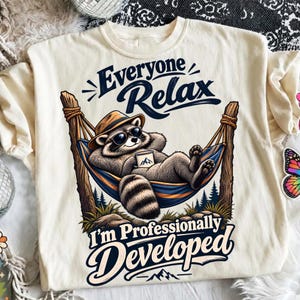 Puede incluir: Una camiseta color crema con el gráfico de un mapache con sombrero y gafas de sol, descansando en una hamaca mientras sostiene una taza. El texto dice Everyone Relax I'm Professionally Developed. El diseño incluye árboles y montañas.