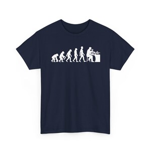 May include: A navy blue short-sleeve t-shirt featuring a white graphic print that depicts the evolutionary progression from a primate to a human sitting at a table playing a card game, with the final figure holding cards and sitting on a stool.