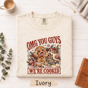 May include: A cream-colored t-shirt featuring a graphic of a distressed gingerbread person with the text "OMG YOU GUYS WE'RE COOKED" in red. The design includes a mixing bowl, a rolling pin, and holiday greenery accents.