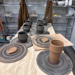 May include: A collection of unfinished ceramic pottery pieces, including several dark gray clay vessels and one tall terracotta-colored cup, each resting on circular wooden bats arranged across a light-colored wooden workbench surface.