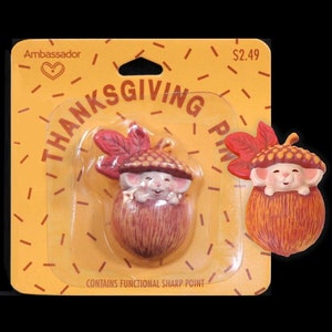 May include: Thanksgiving pin featuring a smiling mouse in an acorn shell, with a red leaf on its head. The pin is on a yellow card with the words "THANKSGIVING PIN" and "Ambassador" printed on it. The card has a heart symbol.