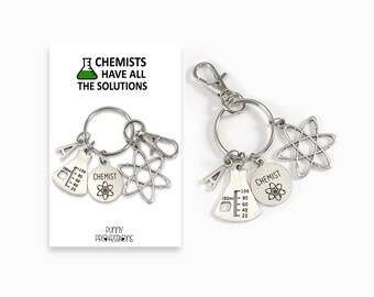May include: Silver keychain with a beaker, atom, and a circle with the word "CHEMIST" and an atom inside. The keychain also has a letter "A" charm. The keychain is attached to a white card with the text "CHEMISTS HAVE ALL THE SOLUTIONS" and a beaker with green liquid.