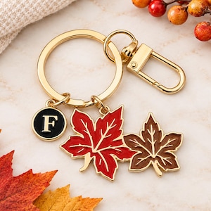 May include: A gold-toned metal keychain featuring a circular ring and a lobster clasp. Attached are three charms: a small black circular charm with the white letter F, a vibrant red maple leaf charm, and a smaller brown maple leaf charm.