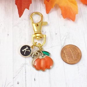 May include: A gold-colored keychain with a lobster clasp, a black circular charm with the letter "K", and a pumpkin charm. A penny is next to the charms. The pumpkin charm is orange with a green stem.