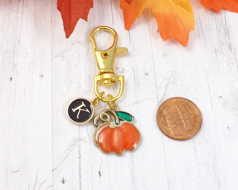 May include: A gold-colored keychain with a lobster clasp, a black circular charm with the letter "K", and a pumpkin charm. A penny is next to the charms. The pumpkin charm is orange with a green stem.