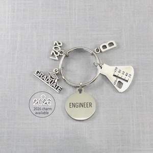 May include: Silver-toned keychain with charms. Charms include a graduation cap, the year "2021", a beaker, and a charm that says "ENGINEER". A smaller charm indicates "2026 charm available".
