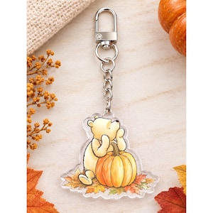May include: A clear acrylic keychain featuring a light yellow bear character resting its chin on its hands while sitting next to a large orange pumpkin. The design is set against a base of colorful autumn leaves and includes a silver metal clasp.