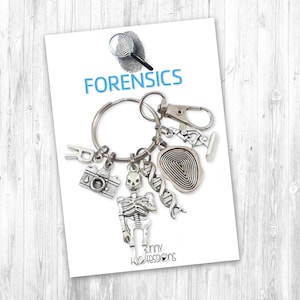 May include: Silver keychain with a skeleton, camera, fingerprint, DNA, and a magnifying glass charm. The keychain has the word "FORENSICS" printed on the card.