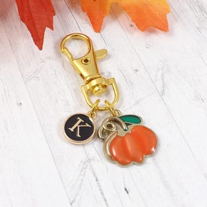 May include: A gold-colored keychain with a lobster clasp, a charm with the letter "K" on a black background, and a charm shaped like an orange pumpkin with a green stem. The keychain is likely a seasonal accessory.