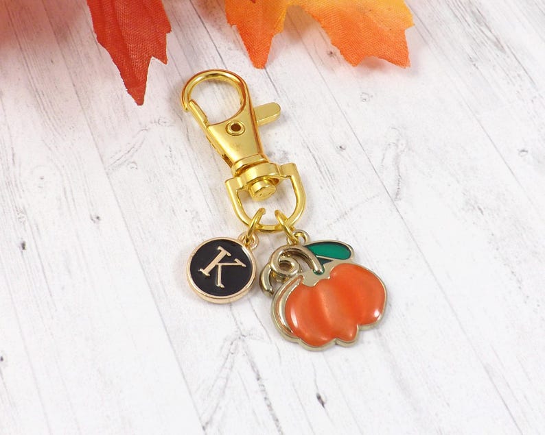 May include: A gold-colored keychain with a lobster clasp, a charm with the letter "K" on a black background, and a charm shaped like an orange pumpkin with a green stem. The keychain is likely a seasonal accessory.