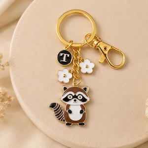 May include: A gold-colored keychain with a raccoon charm, a black letter "T" charm, and white flower charms. The raccoon charm is brown, white, and black. The keychain has a lobster clasp and a key ring.