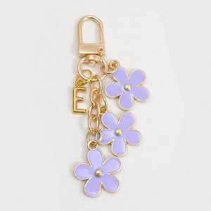 May include: Gold-tone metal keychain with three lavender flower charms and the letter "E". The flowers have a gold outline and a small gold center. The keychain has a lobster clasp.