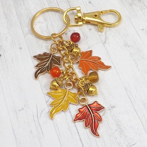 May include: A gold-colored keychain with a lobster clasp and a key ring. The charm features colorful enamel leaves in shades of brown, orange, yellow, and red, along with gold acorns and orange beads. The keychain is designed with an autumn theme.
