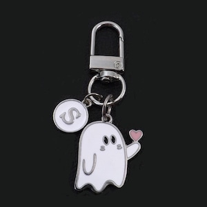 May include: A silver keychain with a white ghost charm holding a pink heart. The keychain also features a round charm with the letter "S" in a white circle. The background is black.