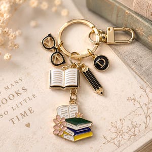 May include: Gold-tone keychain with book-themed charms, including glasses, an open book, a pencil, and a stack of books with flowers. The keychain is on a book with the text "SO MANY BOOKS SO LITTLE TIME".