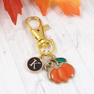 May include: A gold-colored keychain with a lobster clasp, a black circular charm with the letter "K", and a charm shaped like an orange pumpkin with a green stem. The pumpkin charm has a silver outline.