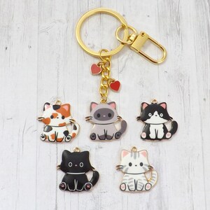 May include: A gold-colored keychain with five cat charms. The charms are in various colors, including calico, gray, black, and white, with heart-shaped accents. The keychain has a clasp and a ring.