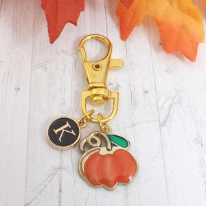 May include: A gold-colored keychain with a lobster clasp, a round charm with the letter "K", and a pumpkin charm. The pumpkin charm is orange with a green leaf and gold trim. The background includes fall leaves.