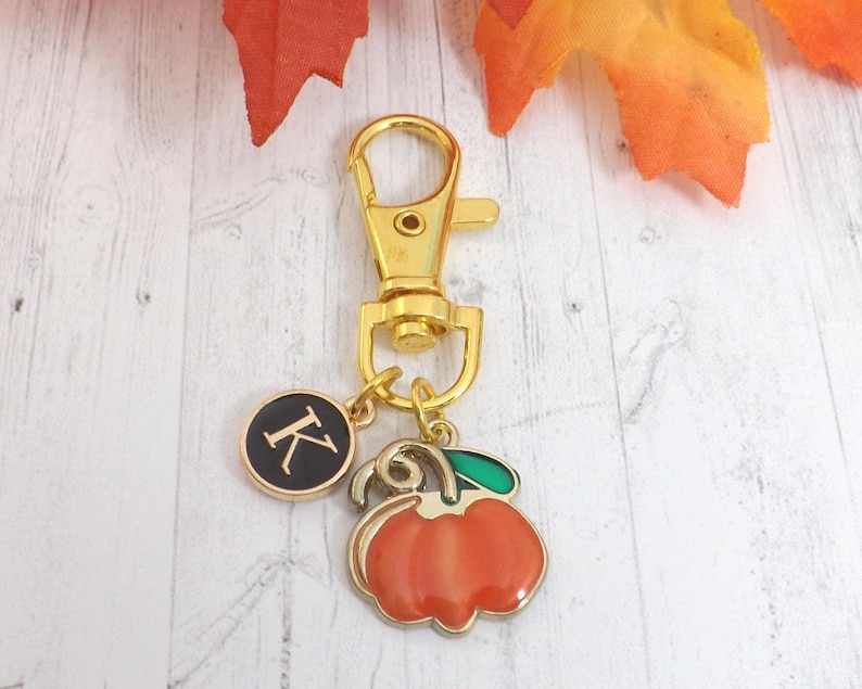 May include: A gold-colored keychain with a lobster clasp, a round charm with the letter "K", and a pumpkin charm. The pumpkin charm is orange with a green leaf and gold trim. The background includes fall leaves.