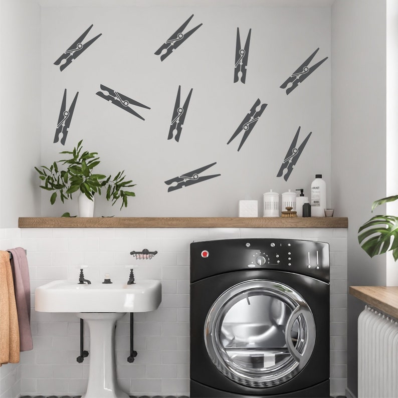 May include: A laundry room scene featuring a black washing machine, a white pedestal sink, and a wooden shelf. The wall is decorated with dark gray clothespin decals. A green plant in a white pot adds a touch of nature.